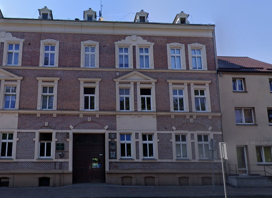 Dwupiętrowa kamienica z cegły z jasnymi ramami okiennymi i ciemnymi, drewnianymi drzwiami wejściowymi. Budynek o symetrycznej fasadzie, z numerem '9' nad wejściem.