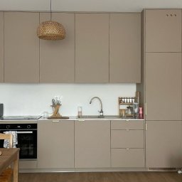 Las u Nas - Beżowa zabudowa kuchenna z drewnianym stołem i lampą w stylu boho. Minimalistyczny design, jasne wnętrze. Widoczny zlew, piekarnik i dodatki kuchenne.