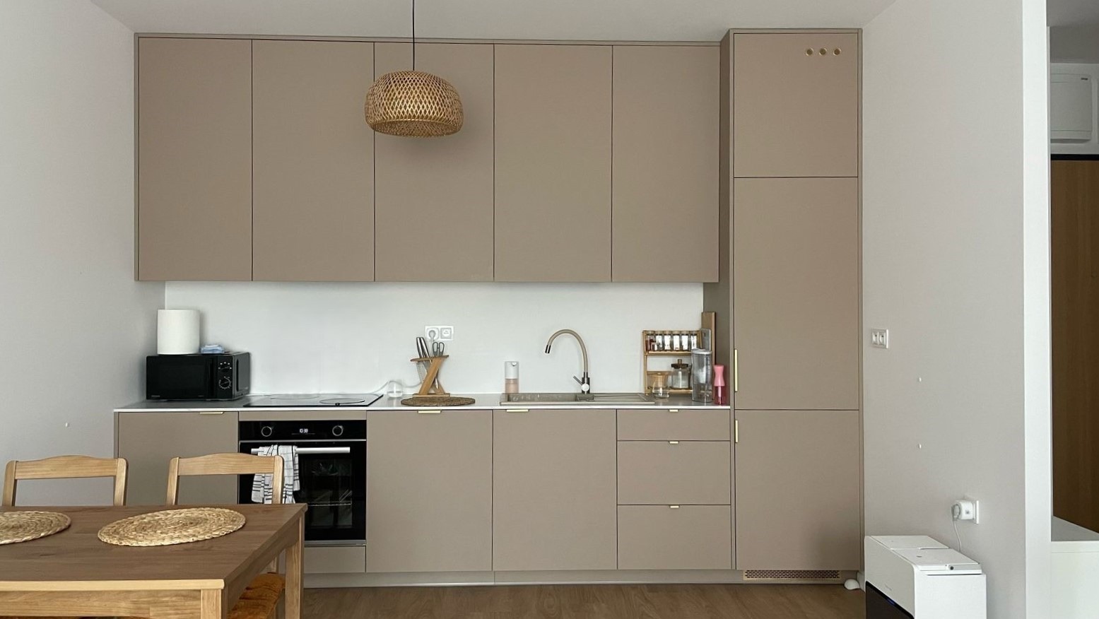 Beżowa zabudowa kuchenna z drewnianym stołem i lampą w stylu boho. Minimalistyczny design, jasne wnętrze. Widoczny zlew, piekarnik i dodatki kuchenne.