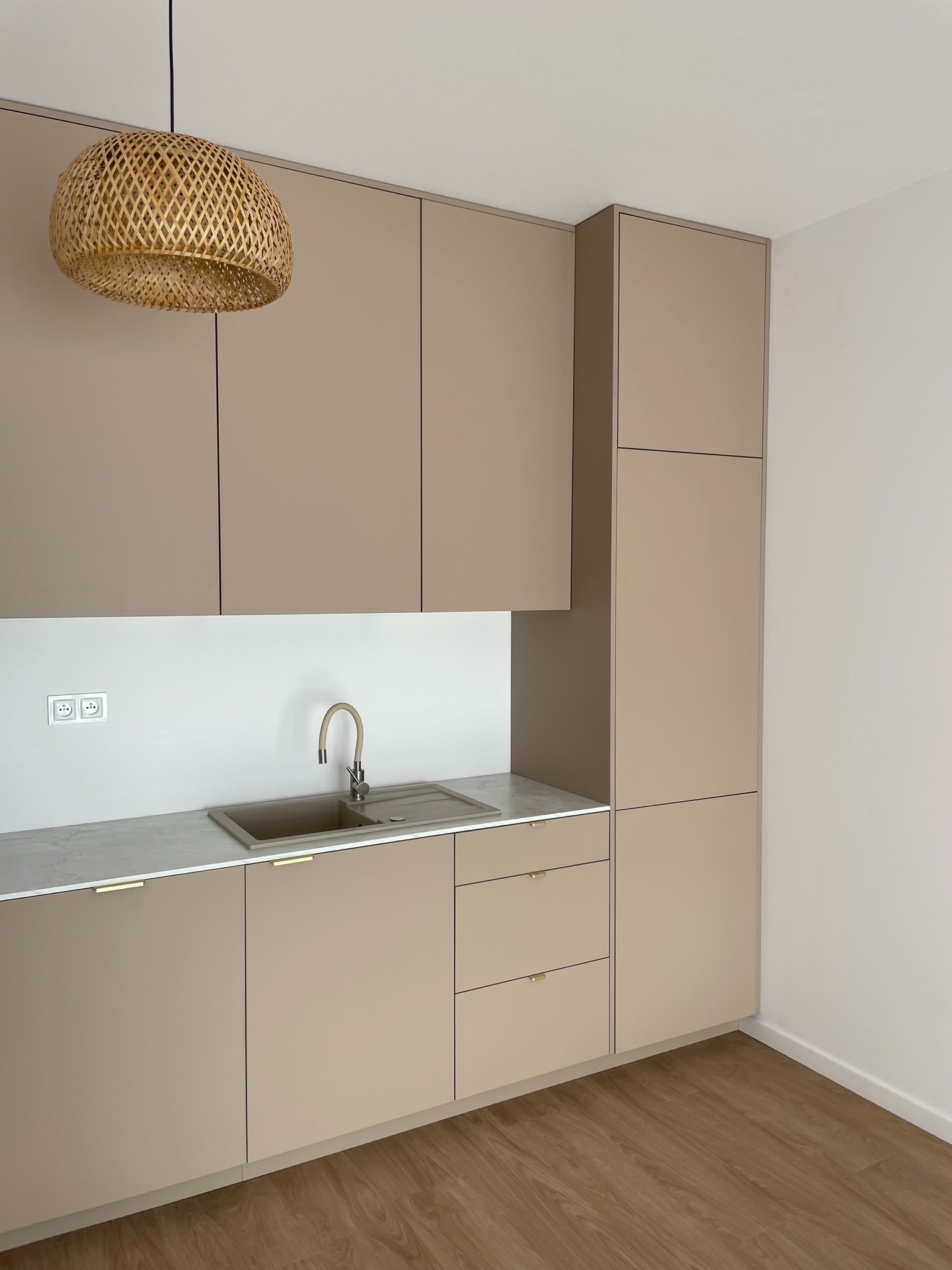 Beżowa kuchnia na wymiar z drewnianą lampą. Umywalka w kolorze zabudowy, minimalistyczne uchwyty. Nowoczesny design, parkiet na podłodze.