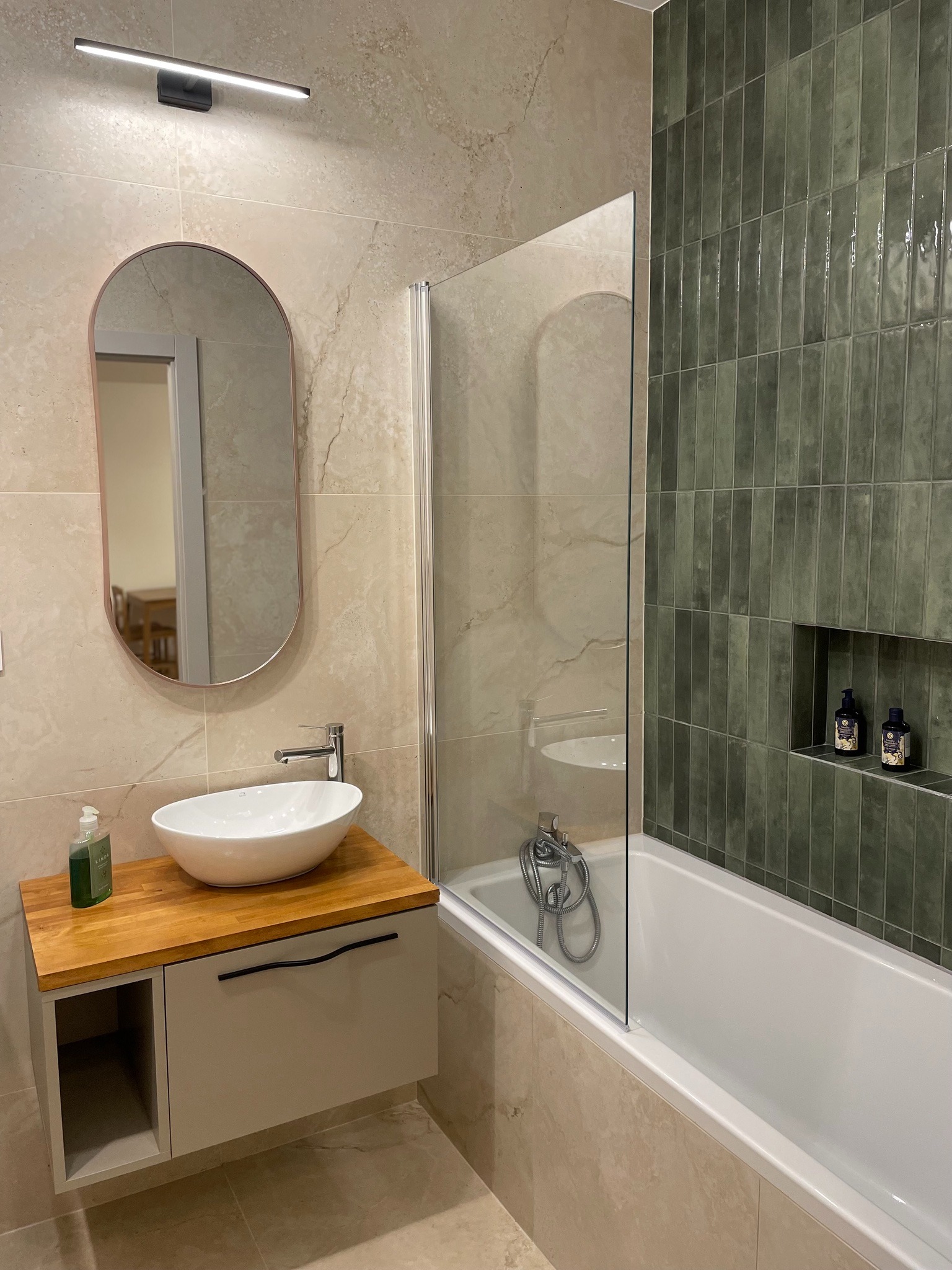 Nowoczesna łazienka z umywalką nablatową na szafce z drewnianym blatem, zielonymi płytkami i wanną z parawanem. W lustrze odbicie pokoju z drewnianym stołem.