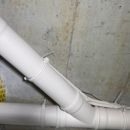 NEXTHOME KOVALEVYCH SPÓŁKA Z OGRANICZONĄ ODPOWIEDZIALNOŚCIĄ - Białe rury wentylacyjne po malowaniu, zabezpieczone taśmą malarską na tle surowego, betonowego stropu. Widoczne przewody elektryczne.