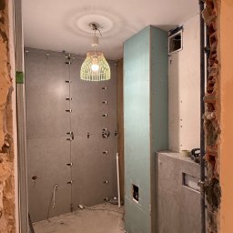 NEXTHOME KOVALEVYCH SPÓŁKA Z OGRANICZONĄ ODPOWIEDZIALNOŚCIĄ - Wnętrze łazienki w trakcie remontu: szare płytki z krzyżykami dystansowymi, zabudowa z płyty gipsowej, widoczne instalacje i surowy stan ścian. W tle lampa z abażurem.