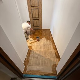 NEXTHOME KOVALEVYCH SPÓŁKA Z OGRANICZONĄ ODPOWIEDZIALNOŚCIĄ - Lakierowanie parkietu w korytarzu. Mężczyzna w białej koszulce aplikuje lakier pędzlem. Widoczna puszka z lakierem i drewniane drzwi w tle.