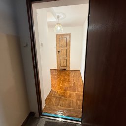 NEXTHOME KOVALEVYCH SPÓŁKA Z OGRANICZONĄ ODPOWIEDZIALNOŚCIĄ - Widok z korytarza przez otwarte, ciemne drzwi na odnowione wnętrze: parkiet, białe ściany, drewniane drzwi i lampa. Akcent na wykończenie podłogi.