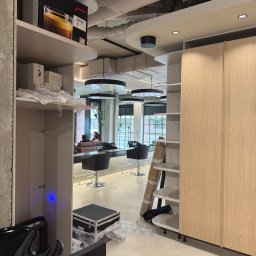 NEXTHOME KOVALEVYCH SPÓŁKA Z OGRANICZONĄ ODPOWIEDZIALNOŚCIĄ - Nowoczesne wnętrze salonu z betonowym sufitem i odkrytymi rurami, wyposażone w drewniane szafki, czarne fotele fryzjerskie i okrągłe lampy wiszące, w trakcie urządzania.