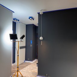 NEXTHOME KOVALEVYCH SPÓŁKA Z OGRANICZONĄ ODPOWIEDZIALNOŚCIĄ - Korytarz w trakcie prac wykończeniowych: ściany w odcieniach ciemnej szarości i bieli, z niebieską taśmą malarską i folią na podłodze. Oświetlenie robocze na statywie.