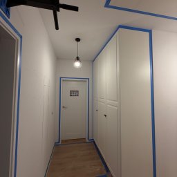 NEXTHOME KOVALEVYCH SPÓŁKA Z OGRANICZONĄ ODPOWIEDZIALNOŚCIĄ - Świeżo pomalowany na biało korytarz z niebieską taśmą malarską na krawędziach, zabezpieczoną podłogą, szafą wnękową, nowoczesną lampą i czarnym drążkiem do podciągania.