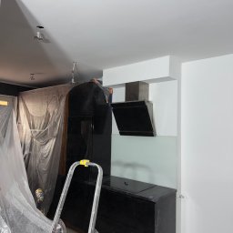 NEXTHOME KOVALEVYCH SPÓŁKA Z OGRANICZONĄ ODPOWIEDZIALNOŚCIĄ - Nowoczesna kuchnia w trakcie prac wykończeniowych: czarne, błyszczące szafki, okap, jasny panel szklany. Świeżo pomalowane białe ściany i sufit, widoczna drabina i folia zabezpieczająca.