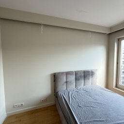 NEXTHOME KOVALEVYCH SPÓŁKA Z OGRANICZONĄ ODPOWIEDZIALNOŚCIĄ - Jasna sypialnia z beżowymi ścianami, białym sufitem z listwą i drewnianą podłogą, z szarym łóżkiem, gniazdkami i wiszącymi przewodami, wskazująca na świeże wykończenie wnętrza.
