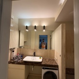 NEXTHOME KOVALEVYCH SPÓŁKA Z OGRANICZONĄ ODPOWIEDZIALNOŚCIĄ - Nowoczesna łazienka z białą umywalką na ciemnym, żyłkowanym blacie, dużym lustrem z trzema czarnymi lampami i pralką Haier. Beżowe płytki, białe szafki z ryflowanym frontem.