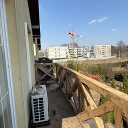 NEXTHOME KOVALEVYCH SPÓŁKA Z OGRANICZONĄ ODPOWIEDZIALNOŚCIĄ - Balkon z masywną drewnianą balustradą w kształcie X, na którym leżą narzędzia obok jednostki klimatyzacji, a w tle widać plac budowy z żurawiem.