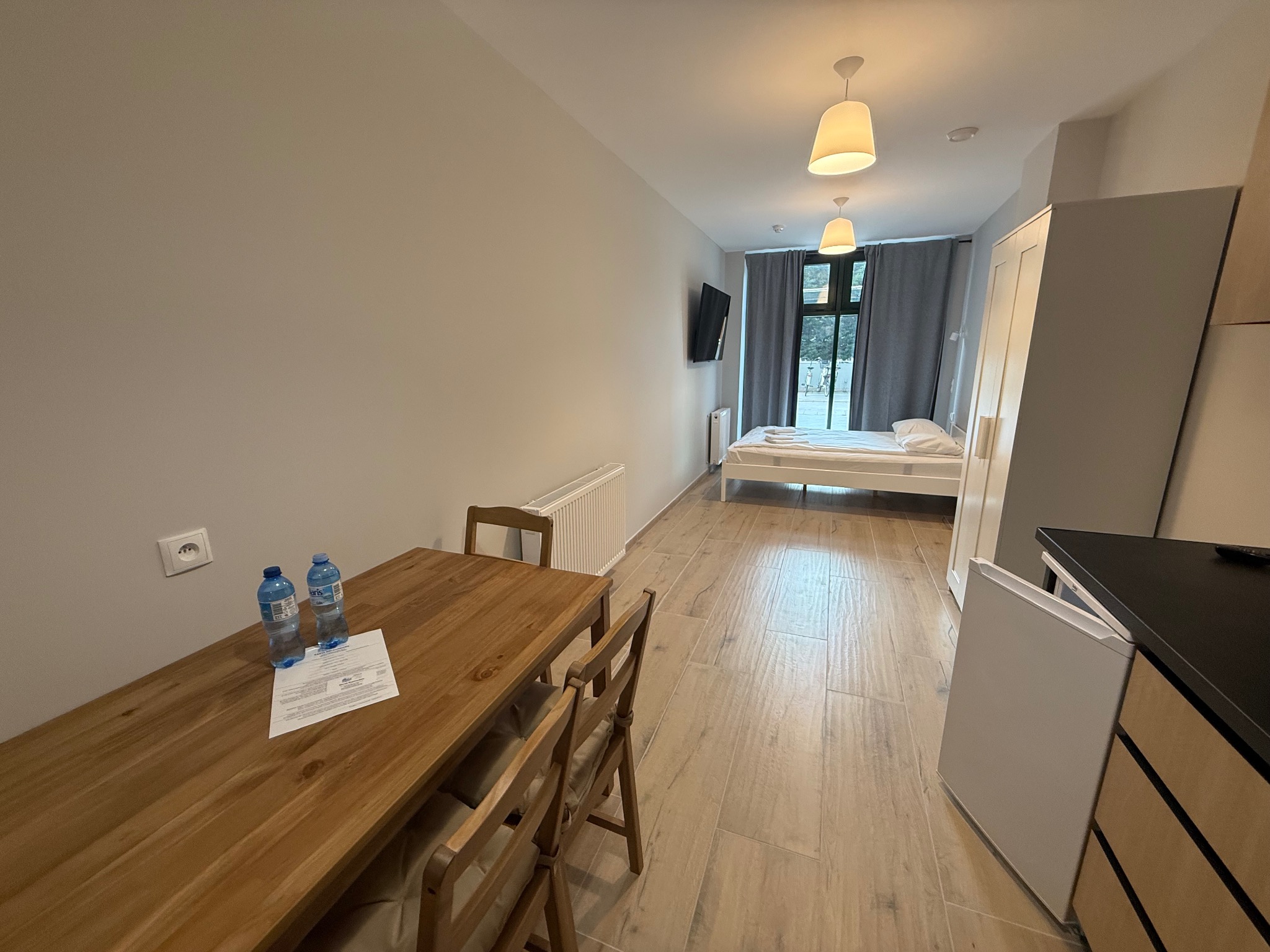 Jasne wnętrze pokoju z drewnianym stołem, krzesłami, łóżkiem i telewizorem. Ściany w odcieniach szarości, podłoga wyłożona płytkami imitującymi drewno. Minimalistyczny wystrój.