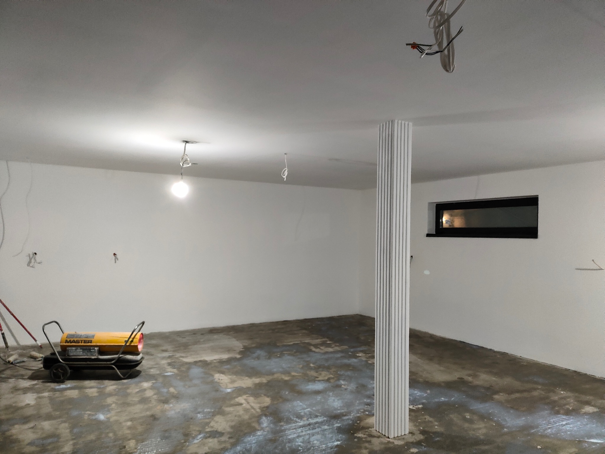 Wnętrze w trakcie prac wykończeniowych: białe ściany, kolumna z płyt karton-gips, widoczny grzejnik MASTER. Podłoga z śladami farby. Wiszące kable.