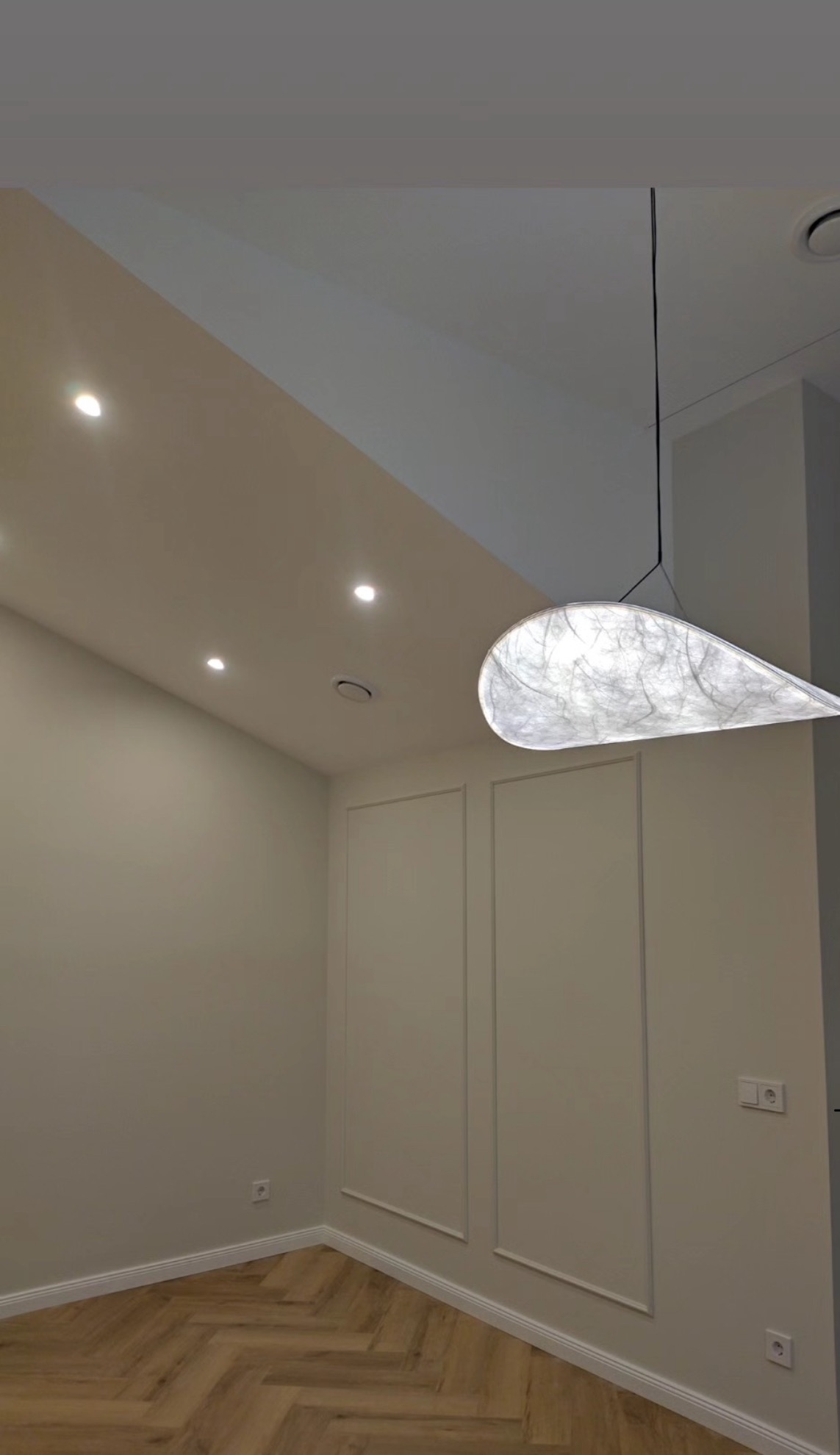 Jasne wnętrze po wykończeniu: parkiet w jodełkę, ściany z dekoracyjnymi listwami, nowoczesna lampa wisząca i punktowe oświetlenie sufitowe.