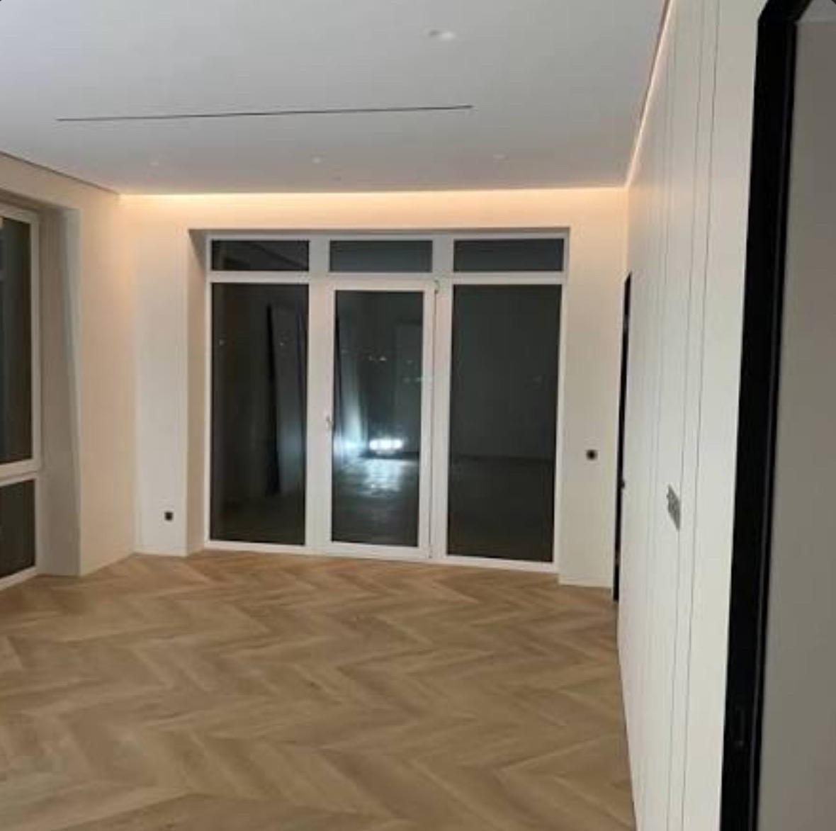 Jasne wnętrze z parkietem ułożonym w jodełkę, duże okna balkonowe z widokiem na nocne miasto, ściany w odcieniach bieli, minimalistyczne oświetlenie sufitowe.