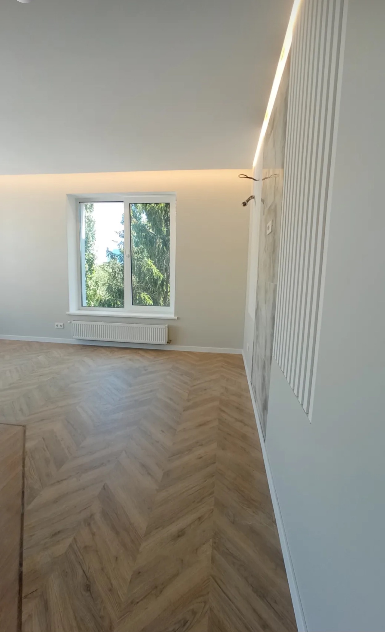 Jasne wnętrze po remoncie: parkiet w jodełkę, białe ściany z dekoracyjnymi panelami i oknem z widokiem na zieleń. Nowoczesne wykończenie, subtelne oświetlenie.