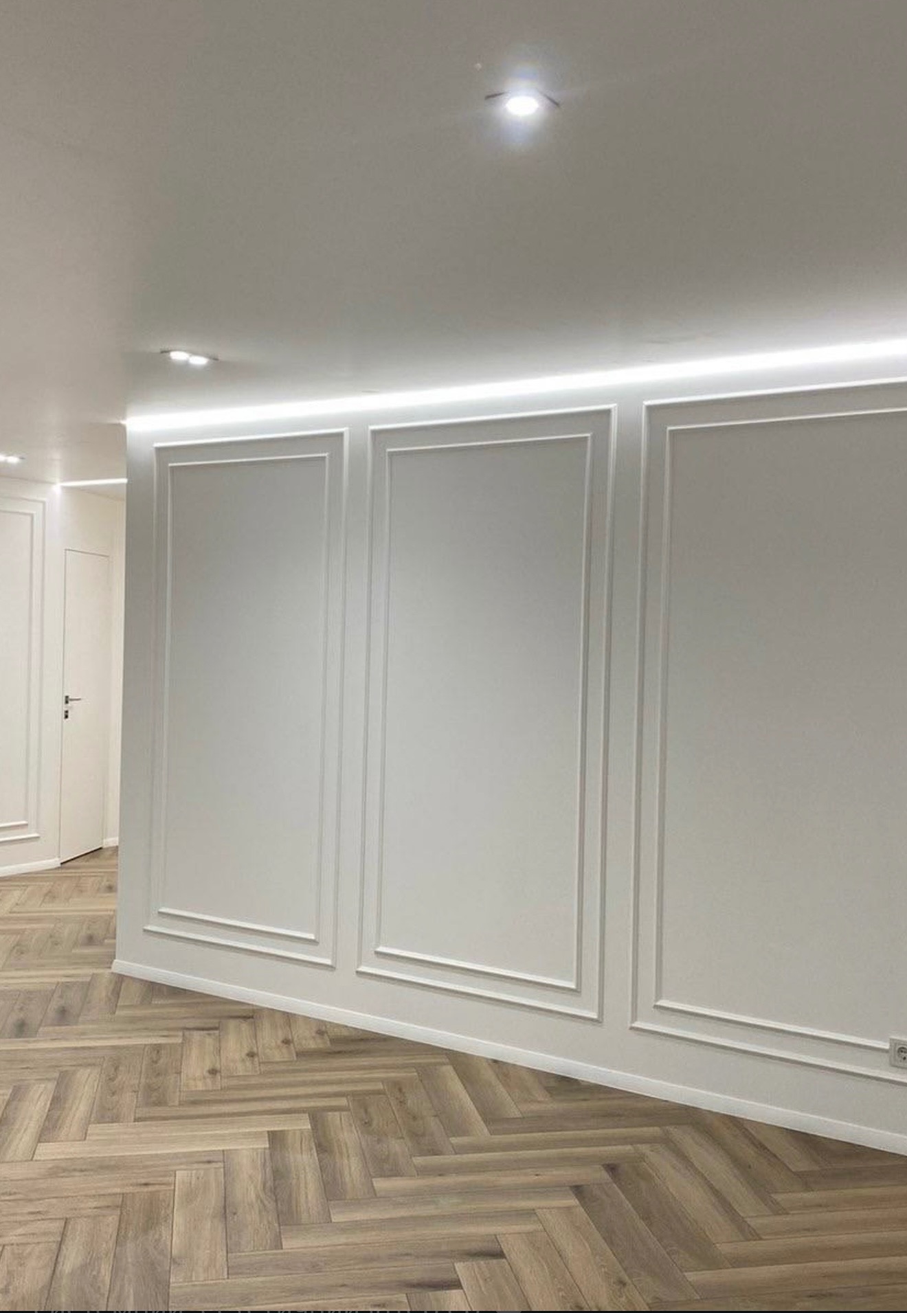 Białe panele ścienne z dekoracyjnymi listwami i oświetleniem LED, parkiet ułożony w jodełkę, widok na drzwi w głębi pomieszczenia, gniazdko elektryczne przy podłodze.