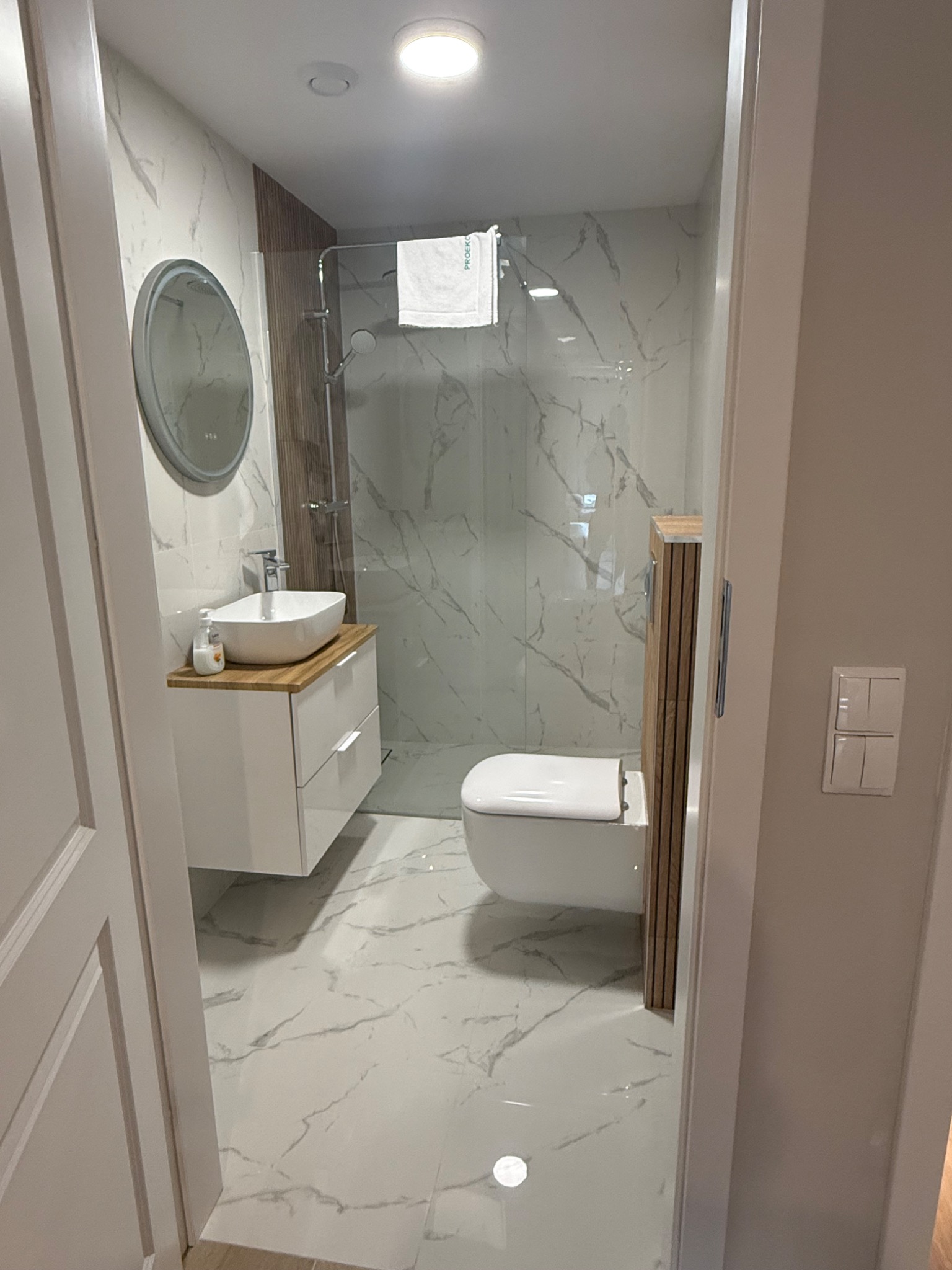 Nowoczesna łazienka z marmurowymi płytkami, białą armaturą i drewnianymi akcentami. Umywalka nablatowa na szafce z szufladami, kabina prysznicowa ze szklaną ścianką.