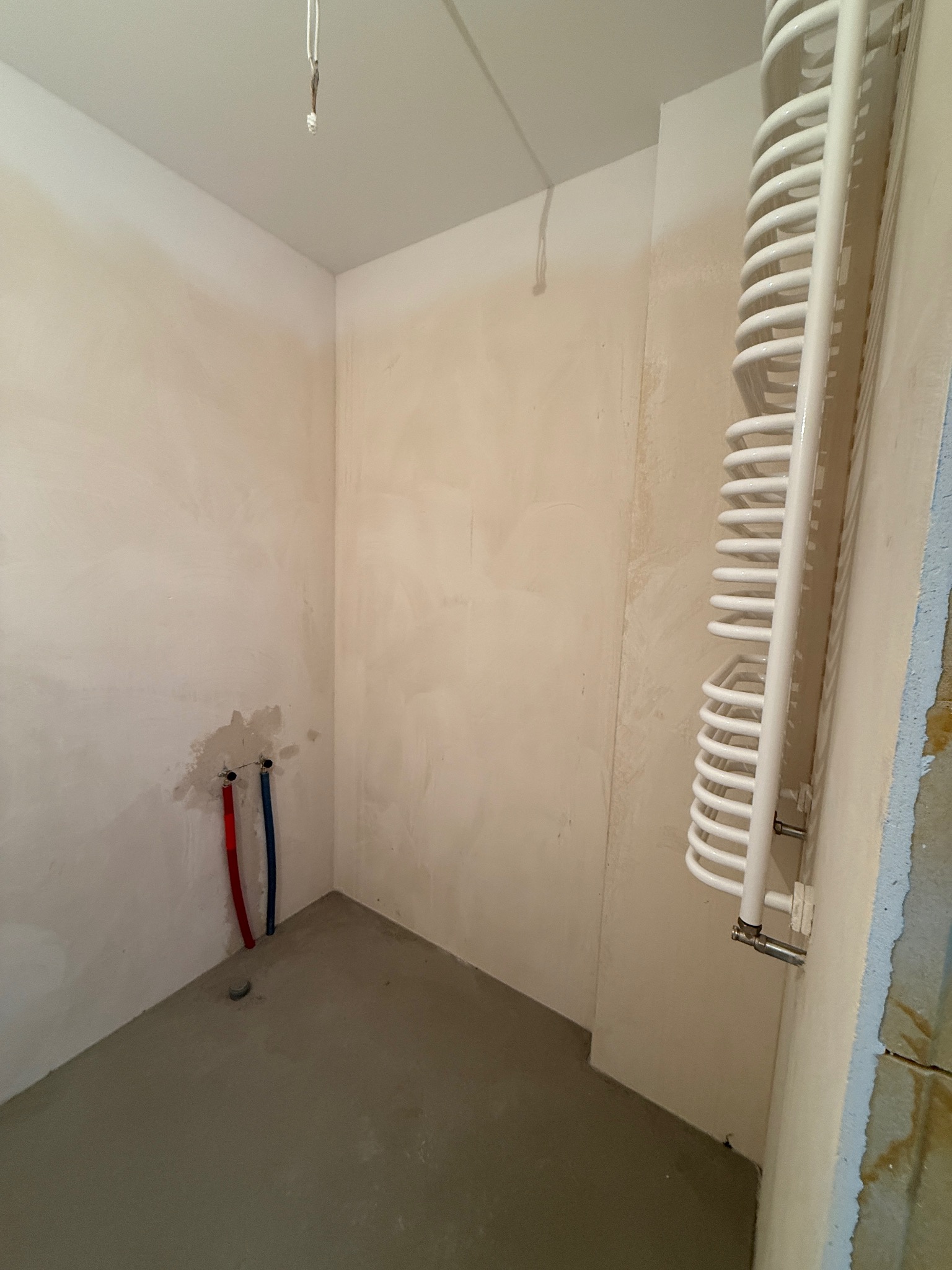 Surowe wnętrze łazienki w trakcie prac: gładzie na ścianach, wylewka na podłodze, zamontowany grzejnik drabinkowy, wystające rury instalacji wodnej.