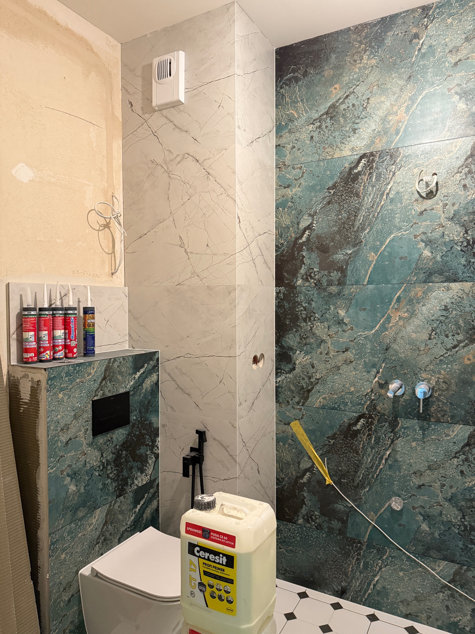 Wnętrze łazienki w trakcie wykańczania: położone płytki imitujące marmur, widoczne materiały budowlane i fragment toalety. Częściowo wykończona ściana.