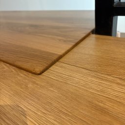 Sklejkovo - Detale wykończenia blatu z jasnego drewna dębowego, z zaokrąglonym rogiem i widoczną strukturą słojów, fragment czarnej nogi stołu w tle.