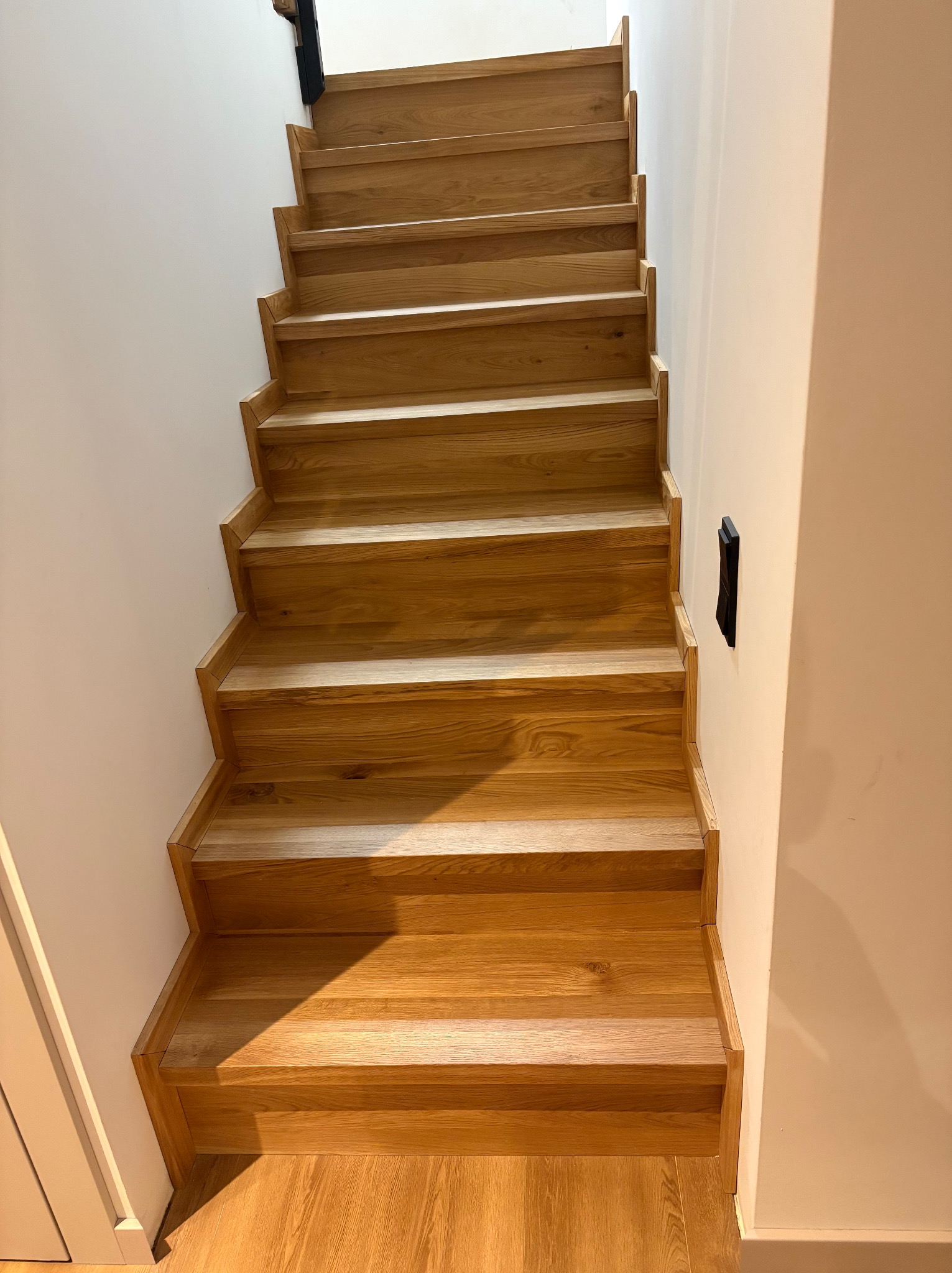 Naturalne, jasne schody z drewna dębowego, widok z dołu. Minimalistyczny design, białe ściany, czarny włącznik. Ciepłe oświetlenie podkreśla strukturę drewna.