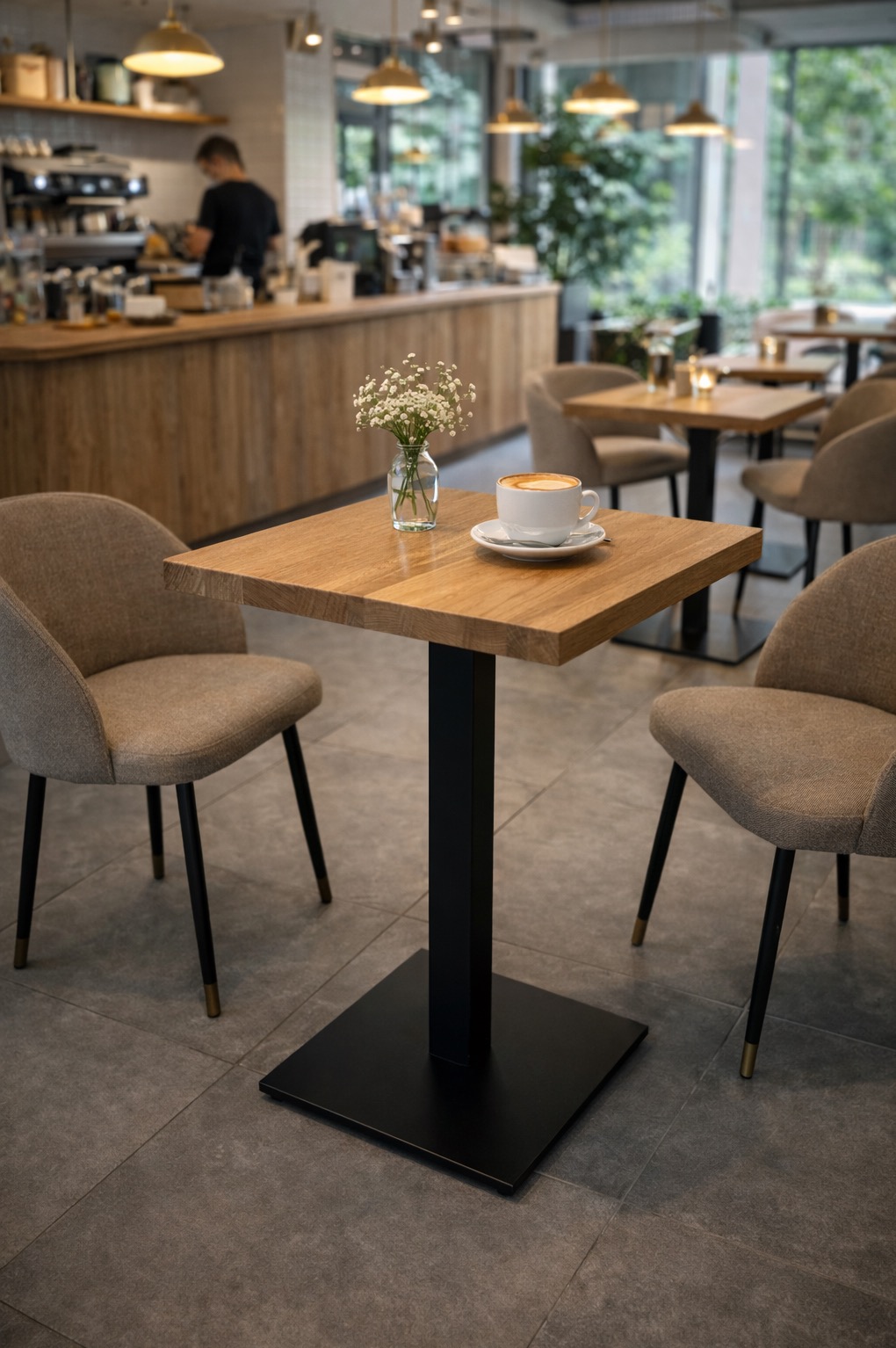 Kwadratowy blat drewniany na czarnej podstawie w kawiarni, z filiżanką kawy i wazonem z kwiatami. Dwa krzesła z beżową tapicerką. W tle bar i pracownik.