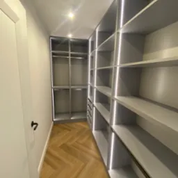 Nowoczesna garderoba z szarymi półkami i oświetleniem LED, parkiet w jodełkę. Widok z perspektywy wejścia, drzwi po lewej stronie.