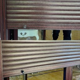 Rodzinny Porządek - Brudne, brązowe żaluzje okienne z widocznymi smugami kurzu. W tle fragment białej komody i czyjeś nogi. Potrzebne sprzątanie.