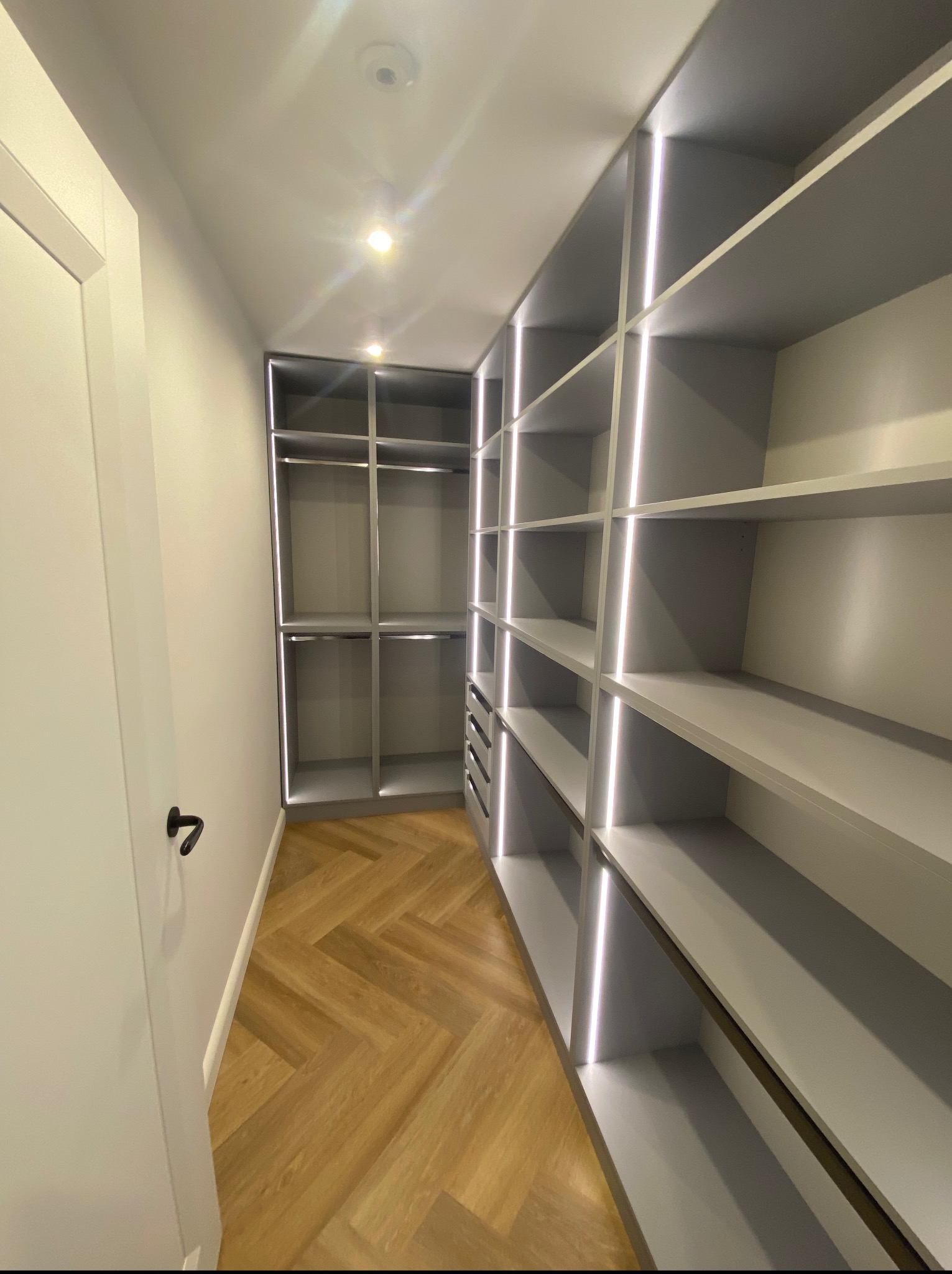 Nowoczesna garderoba z szarymi półkami i oświetleniem LED, parkiet w jodełkę. Widok z perspektywy wejścia, drzwi po lewej stronie.