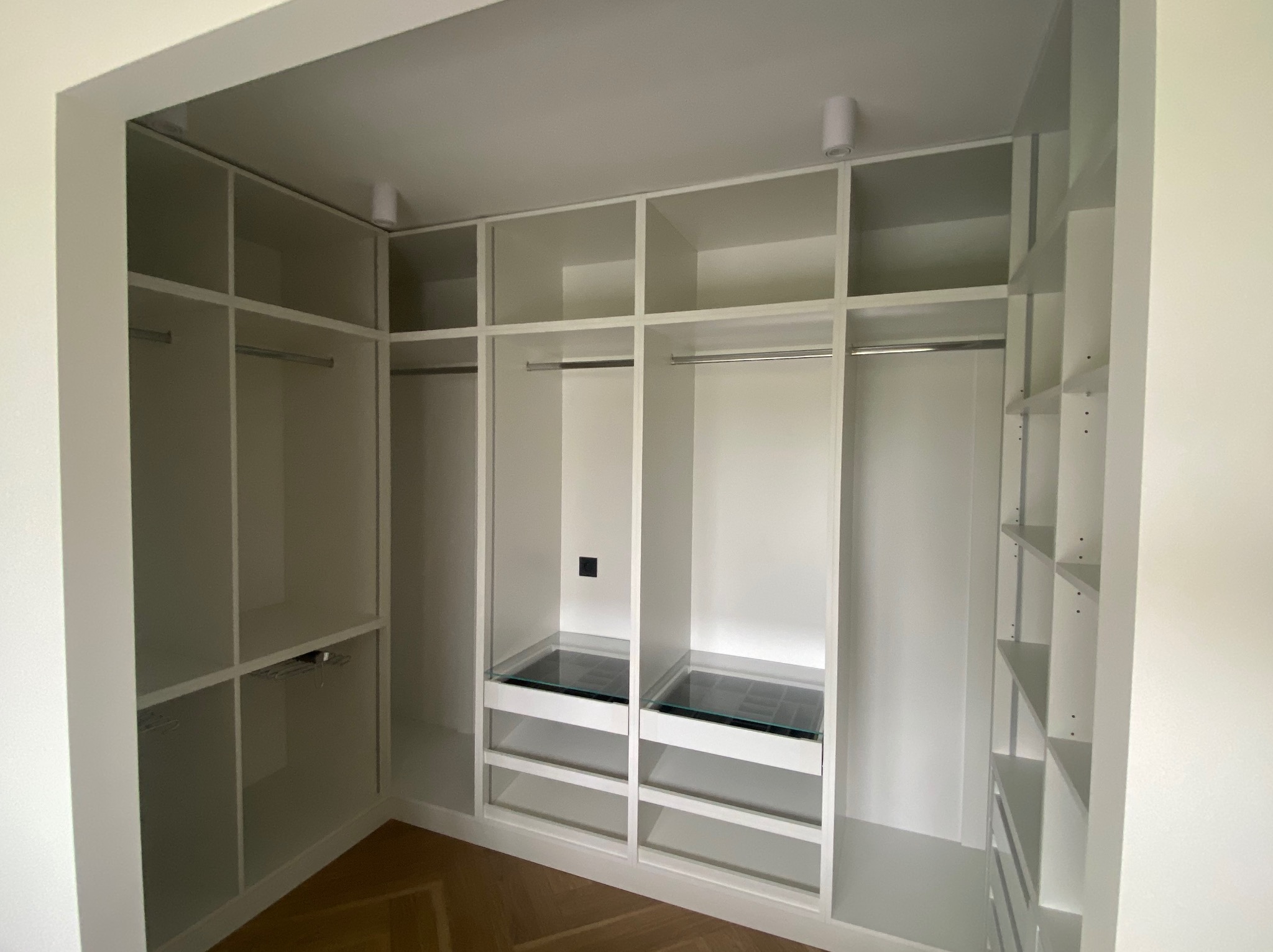 Biała, pusta garderoba w zabudowie z półkami, drążkami i szklanymi elementami. Widoczna drewniana podłoga i oświetlenie punktowe.