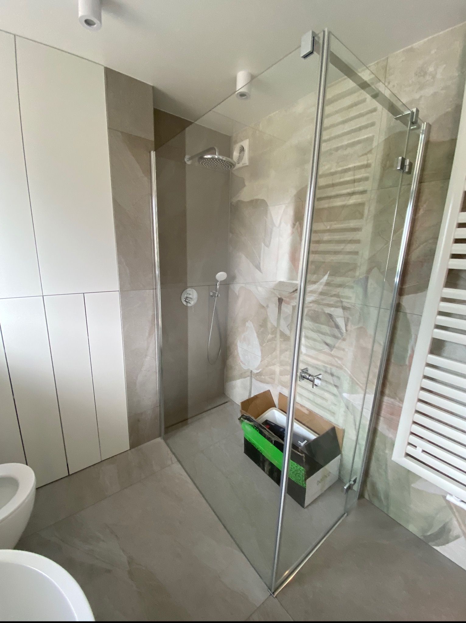 Nowoczesna kabina prysznicowa ze szklanymi drzwiami w wykończonej łazience z szarymi płytkami i białymi meblami. Widoczny grzejnik drabinkowy i karton po produkcie.