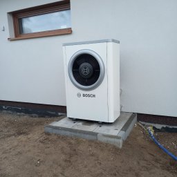 Zakład Instalacyjno-sanitarny INSTAL-MONT Krzysztof Sworski - Biała pompa ciepła Bosch na betonowej podstawie przy ścianie budynku z oknem. Widoczny fragment niebieskiego węża na ziemi.