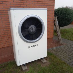 Zakład Instalacyjno-sanitarny INSTAL-MONT Krzysztof Sworski - Biała pompa ciepła Bosch na zewnątrz budynku z cegły, z drabinką opartą o ścianę i krzewem w tle. Widoczne detale montażu na betonowych podstawach.