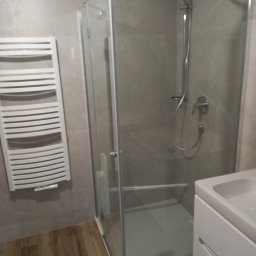 Zakład Instalacyjno-sanitarny INSTAL-MONT Krzysztof Sworski - Nowoczesna kabina prysznicowa z grzejnikiem drabinkowym w łazience wykończonej w szarościach i brązach. Widoczna armatura i umywalka z szafką.