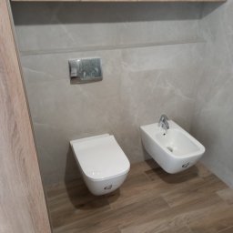 Zakład Instalacyjno-sanitarny INSTAL-MONT Krzysztof Sworski - Nowoczesna łazienka z podwieszaną miską WC i bidetem, wykończona w jasnych odcieniach drewna i szarości. Widoczne płytki imitujące drewno na podłodze.