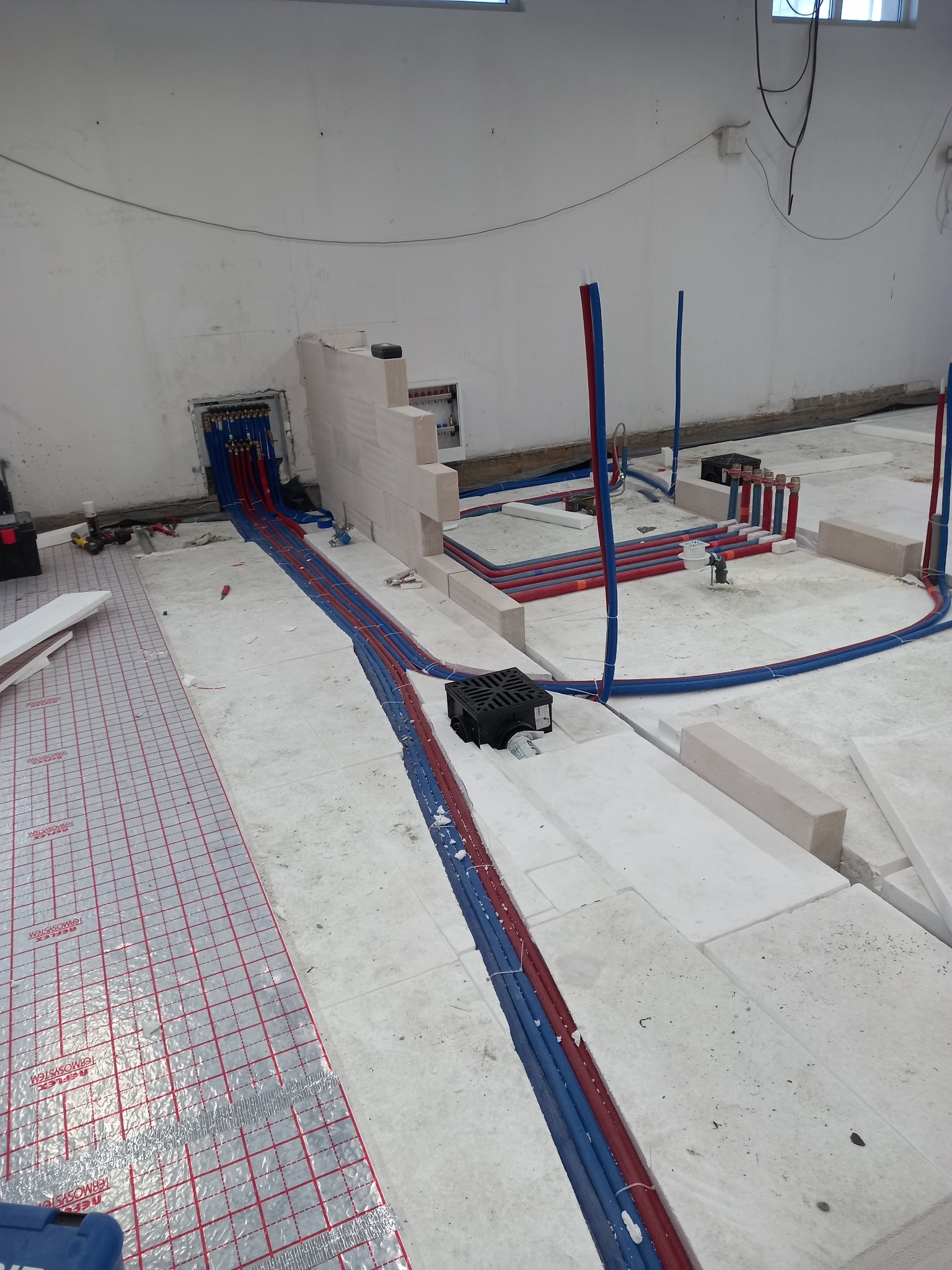 Wnętrze w trakcie instalacji ogrzewania podłogowego: rozłożona mata grzewcza, rury w izolacji termicznej, białe bloczki i rozdzielacz na ścianie.