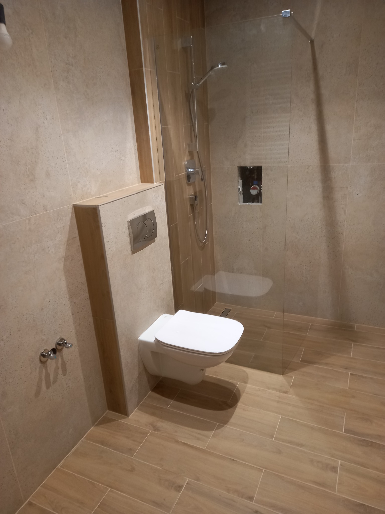 Nowoczesna łazienka z beżowymi płytkami imitującymi drewno, podwieszana toaleta z chromowanym przyciskiem i kabina prysznicowa typu walk-in z odpływem liniowym.