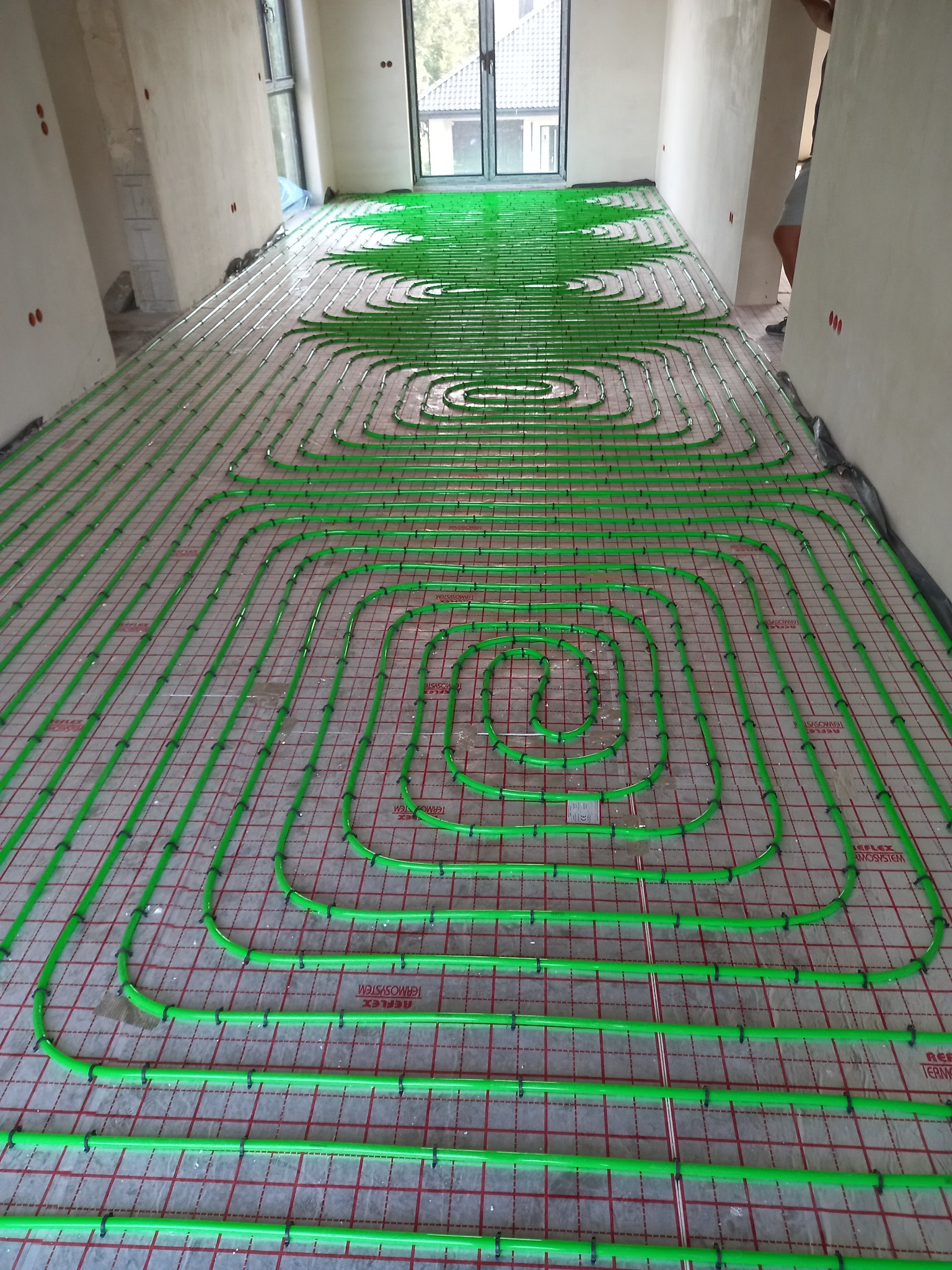 Zielone rury ogrzewania podłogowego, ułożone w pętle na czerwonej siatce, w trakcie montażu w Nakle nad Notecią. Widok z góry na surową podłogę.