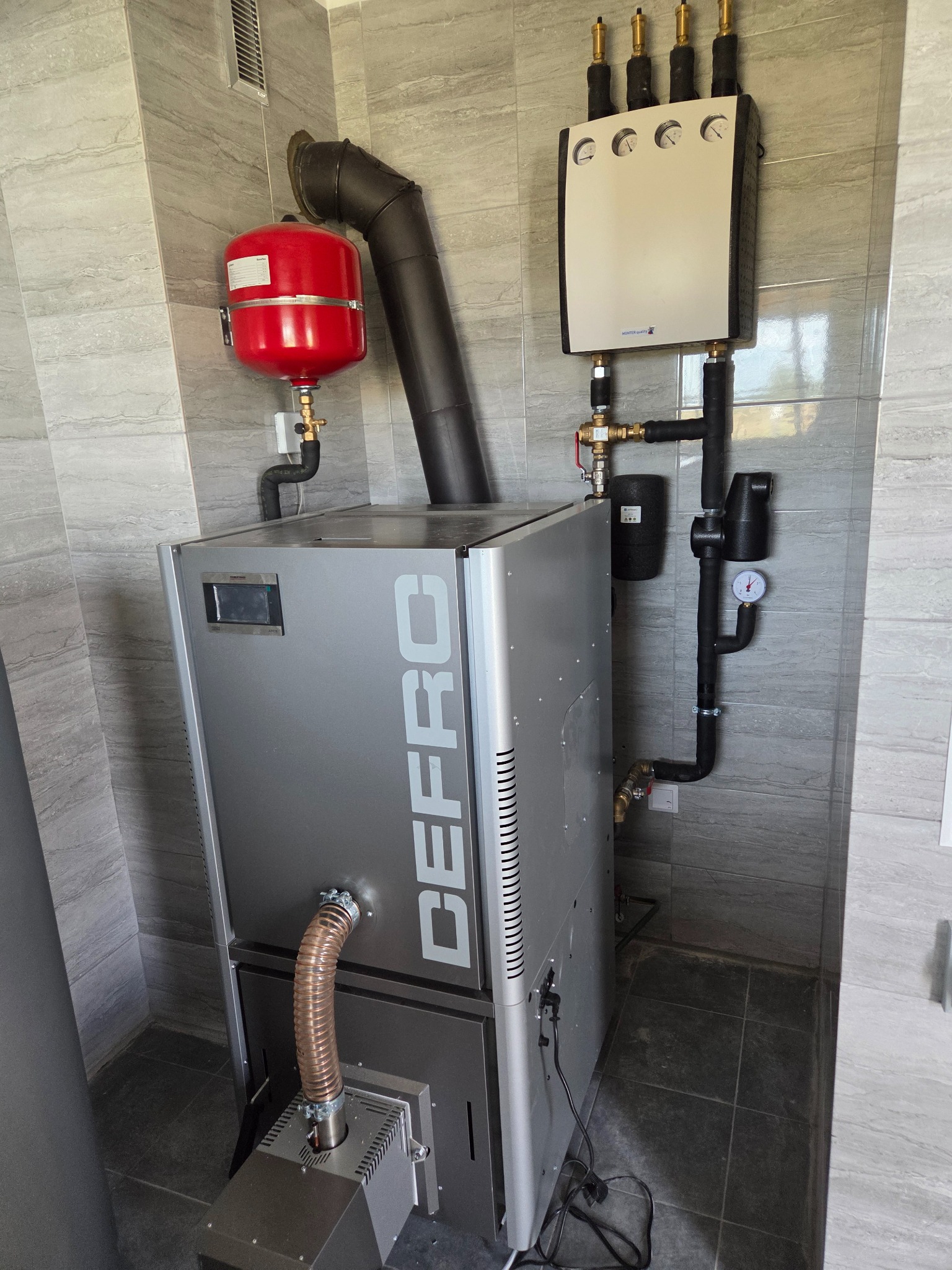 Nowoczesny piec CO marki DEFRO z czerwonym zbiornikiem wyrównawczym i czarną instalacją, zamontowany w pomieszczeniu z szarymi płytkami imitującymi kamień.