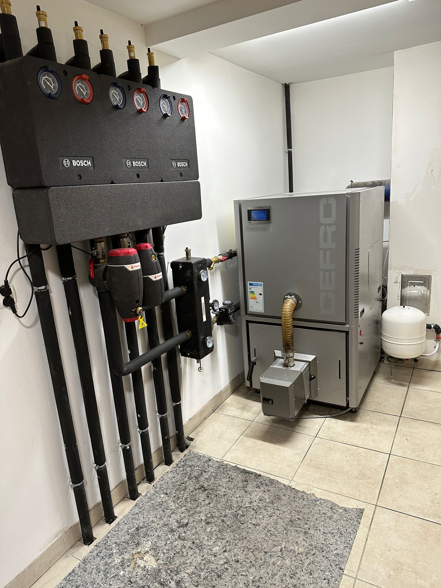 Szary piec CO marki Defro z podłączeniami, czarne rury instalacji grzewczej, szara mata na podłodze w kotłowni. Widok na kompletną instalację.