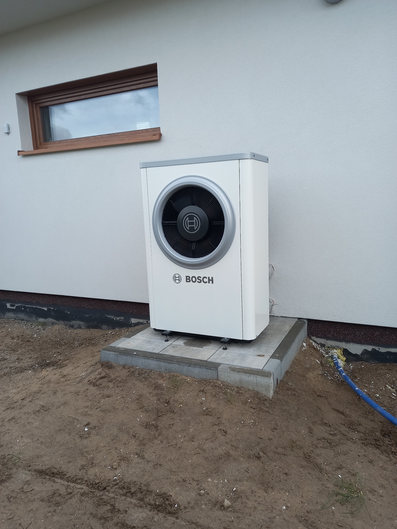 Biała pompa ciepła Bosch na betonowej podstawie przy ścianie budynku z oknem. Widoczny fragment niebieskiego węża na ziemi.