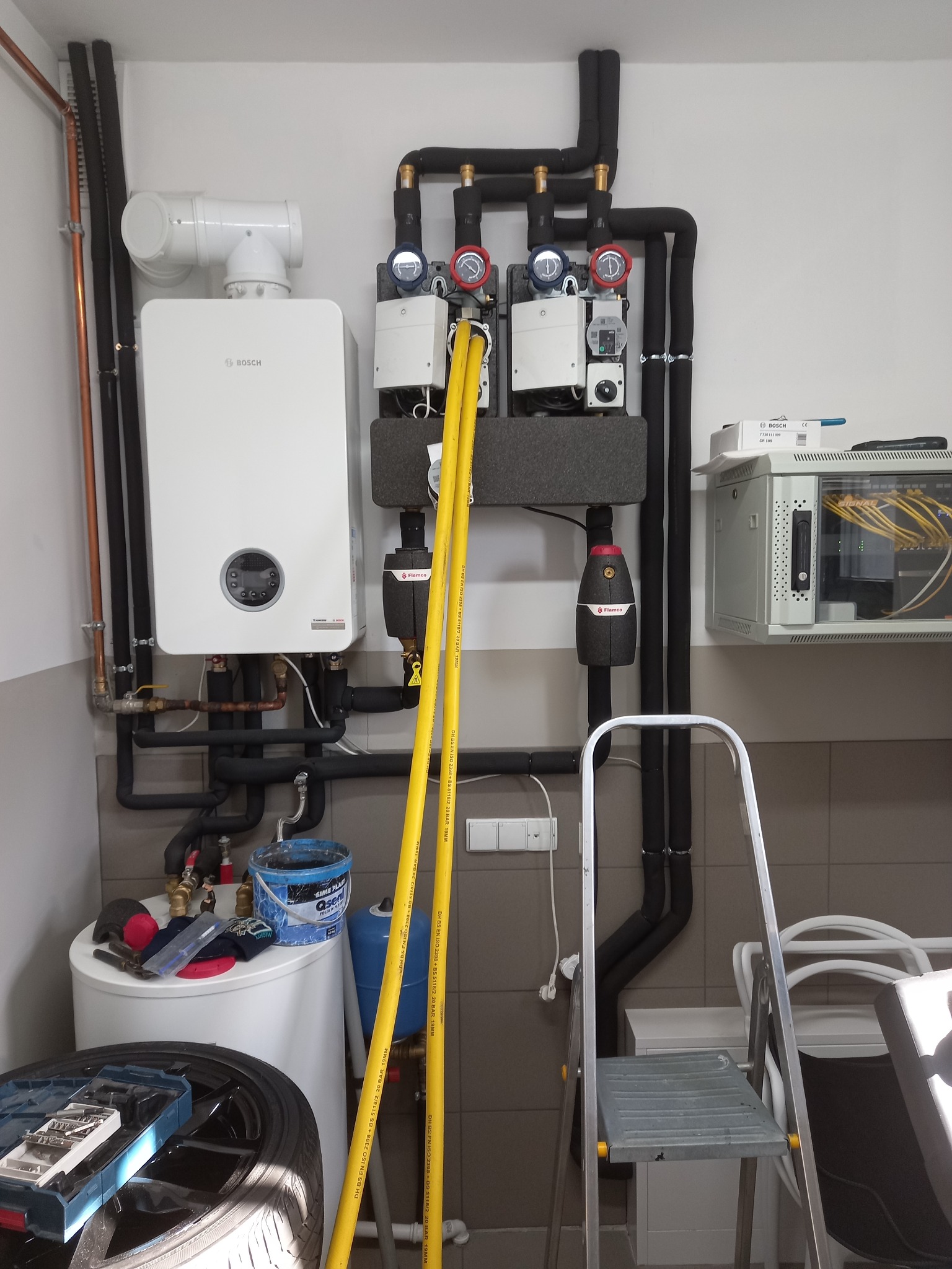 Nowoczesny piec gazowy Bosch z podłączeniami, widoczne rury w izolacji, manometry i elementy instalacji. Na podłodze narzędzia i akcesoria hydrauliczne.