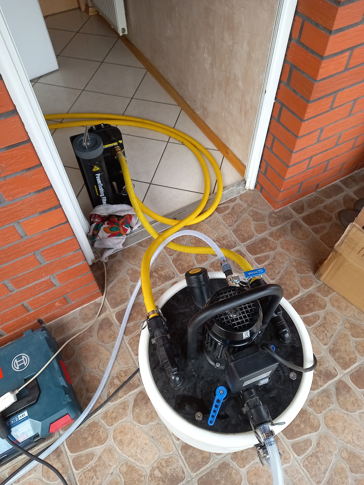 Profesjonalna maszyna do płukania instalacji CO z podłączonymi przewodami, widoczna na tle kafelków i fragmentu ściany z cegły.