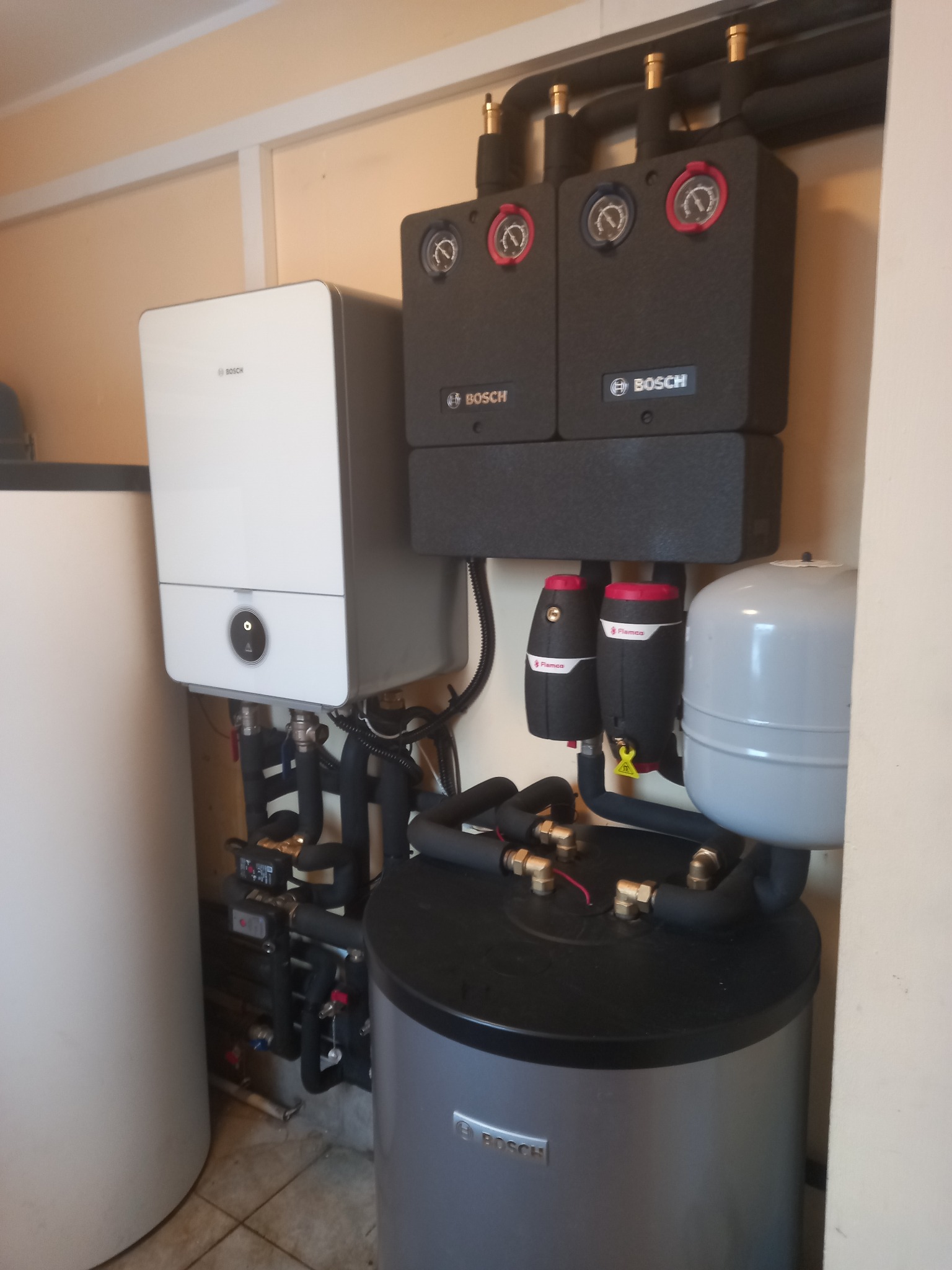 Złożona instalacja grzewcza Bosch z piecem, zasobnikiem i osprzętem, widoczna w pomieszczeniu technicznym. Rury i zawory tworzą skomplikowany system połączeń.