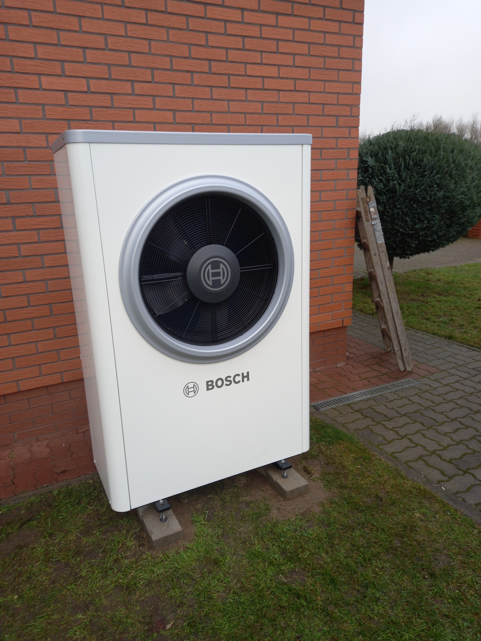 Biała pompa ciepła Bosch na zewnątrz budynku z cegły, z drabinką opartą o ścianę i krzewem w tle. Widoczne detale montażu na betonowych podstawach.
