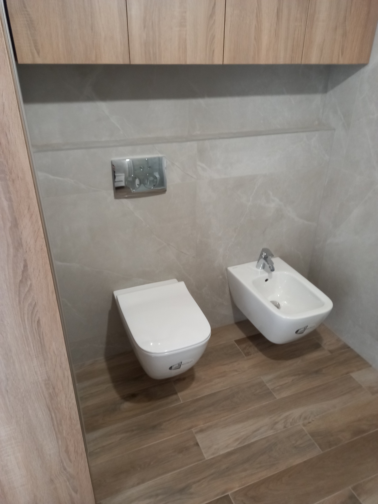 Nowoczesna łazienka z podwieszaną miską WC i bidetem, wykończona w jasnych odcieniach drewna i szarości. Widoczne płytki imitujące drewno na podłodze.