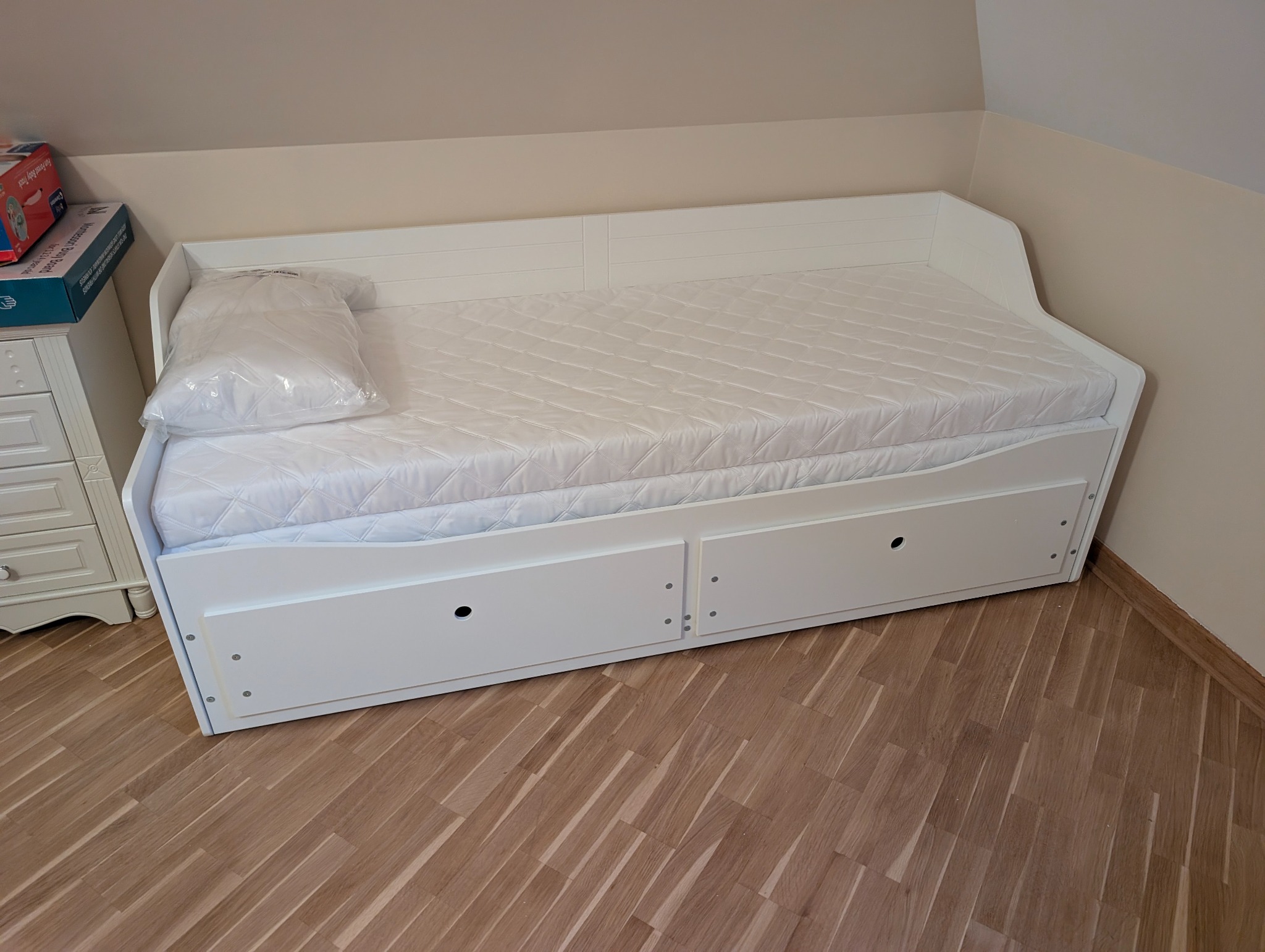 Biała, rozłożona sofa z materacem i szufladami, ustawiona na drewnianej podłodze w rogu pokoju. Poduszka w folii leży na materacu. Widoczny fragment komody.