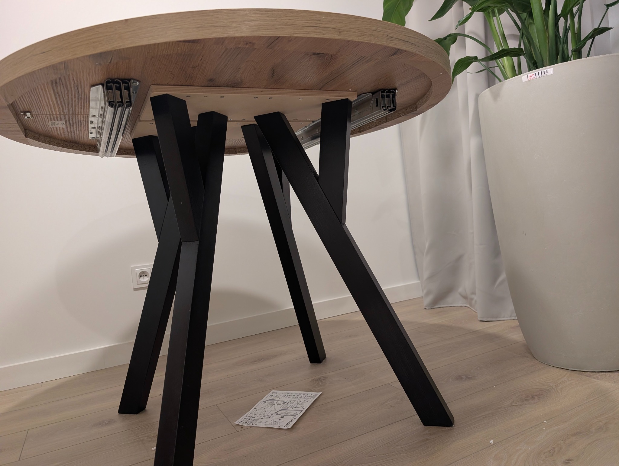 Stół z drewnianym blatem i czarnymi nogami, widok od spodu, instrukcja leży na podłodze. W tle donica z rośliną i biała ściana z gniazdkiem.